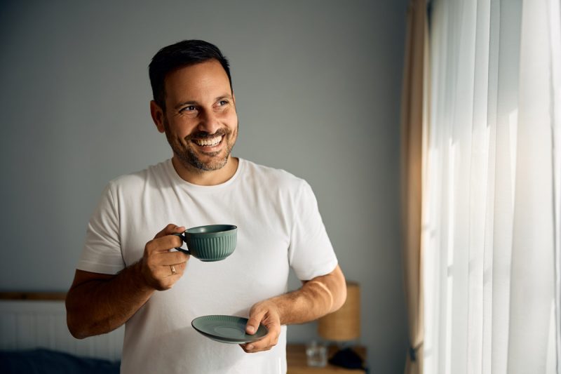 Smiling man enjoying in cup of morning coffee in bedroom. Copy space.